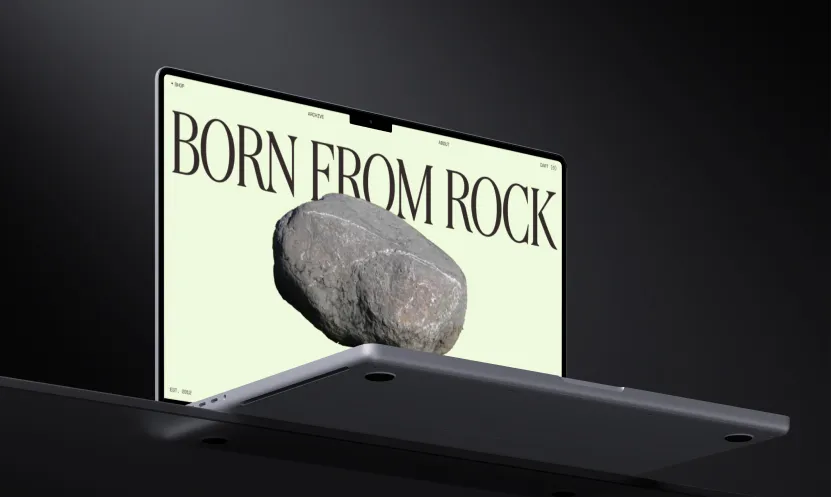 Laptop showing a website with large text 'BORN FROM ROCK' and an image of a gray rock on the screen.