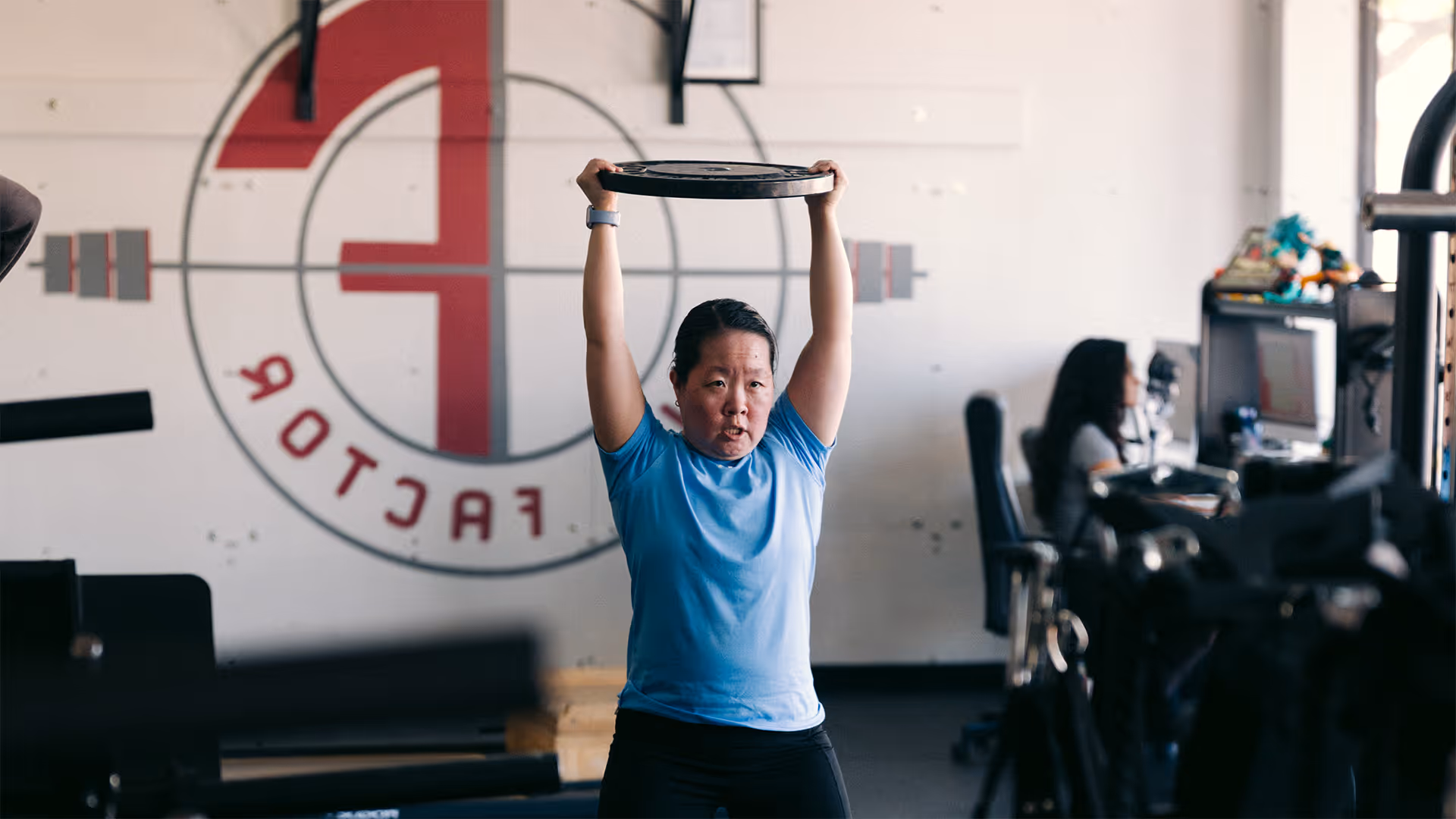 Woman in blue shirt lifting a weight plate overhead inside a gym with fitness equipment and a large 'F' logo on the wall.