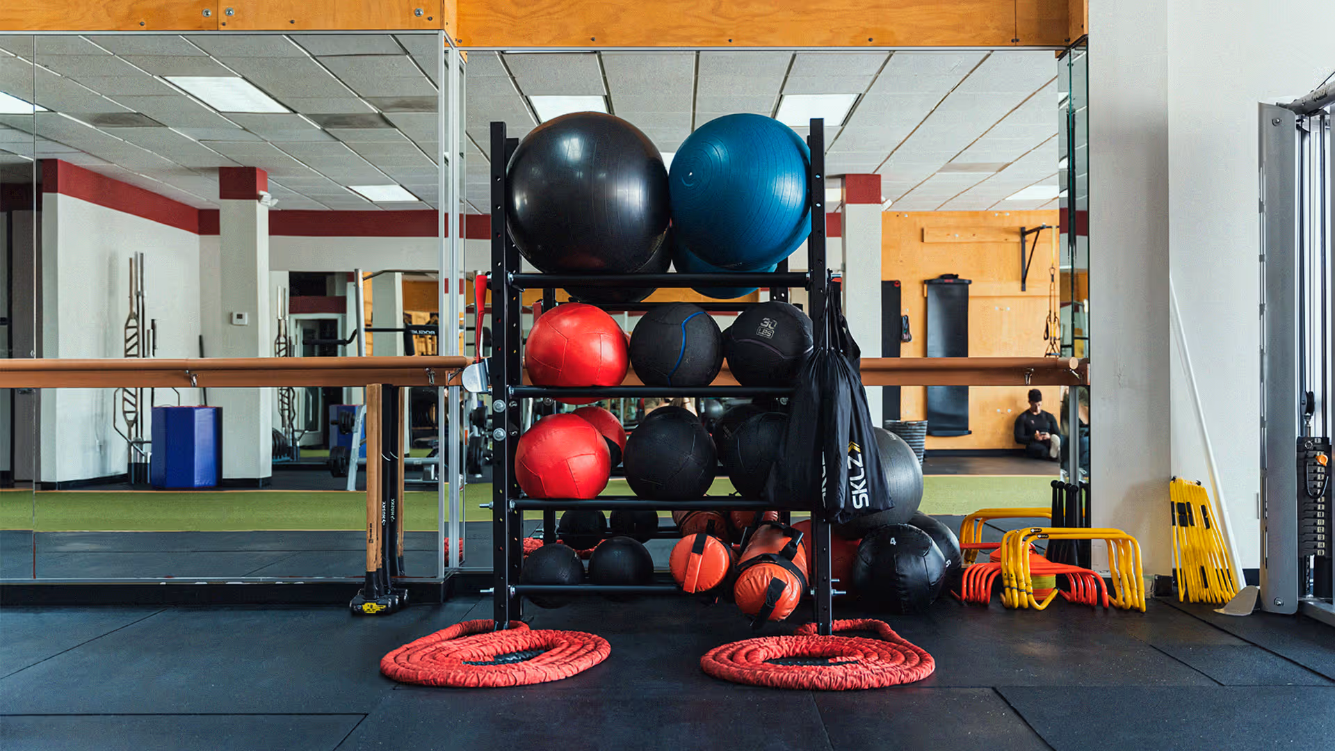 Gym storage rack inside Fit Factor Memorial City with various exercise balls, medicine balls, resistance ropes, and agility hurdles on a black floor with mirrored walls behind.