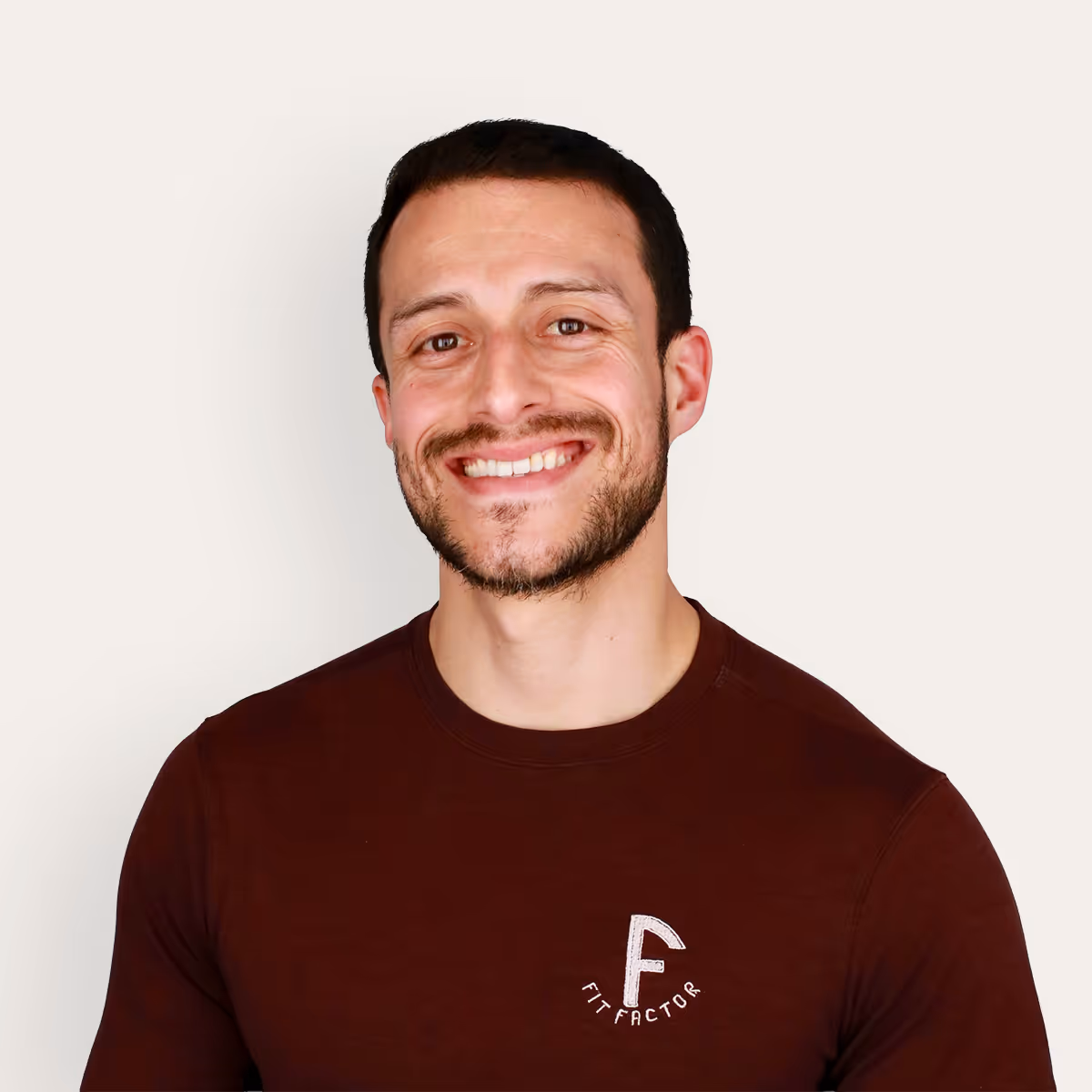 Headshot of Ivan Salazar, Owner & Master Trainer, smiling with short dark hair and beard wearing a maroon 'Fit Factor' shirt against a light background.