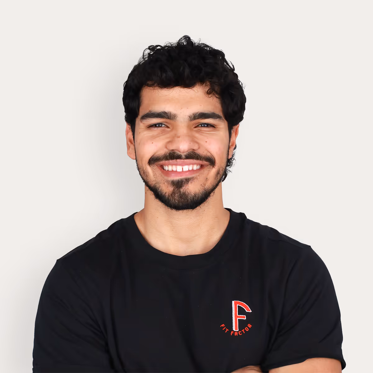 Headshot of Juan Gomez, Certified Personal Trainer, smiling with curly black hair and beard, wearing a black 'Fit Factor' t-shirt, standing against a plain light background.