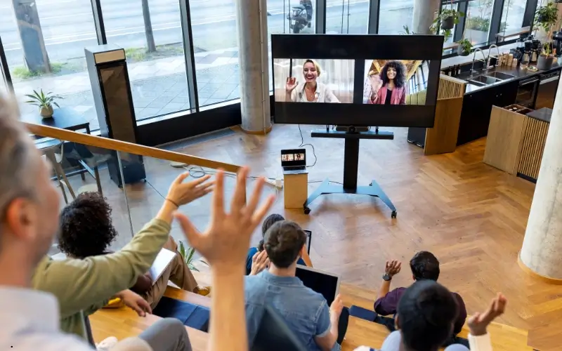 A group of people in a modern workspace interacting with colleagues on a large video screen, representing hybrid event gamification that connects in-person and virtual participants.