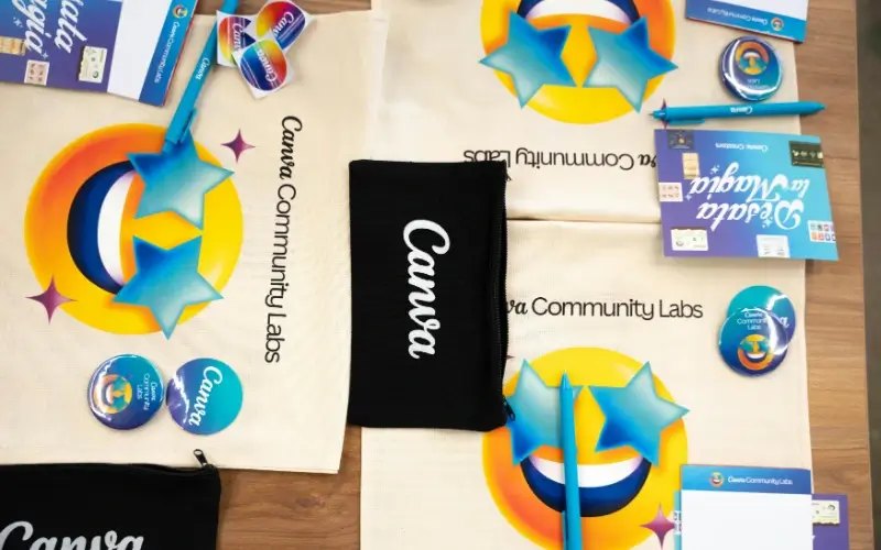 A variety of branded promotional swag for job candidates, including "Canva Community Labs" tote bags, blue pens, stickers, and black zippered pouches.