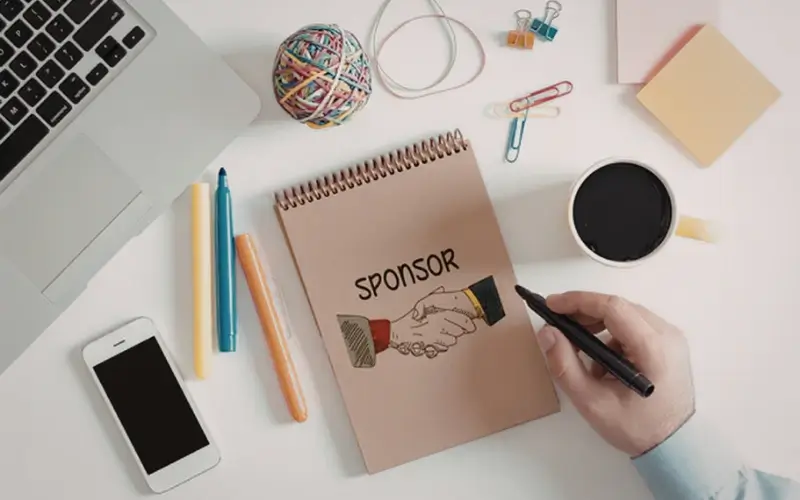 Notebook with the word sponsor and handshake illustration on a desk, representing B2B event sponsorship planning and partner relationships.