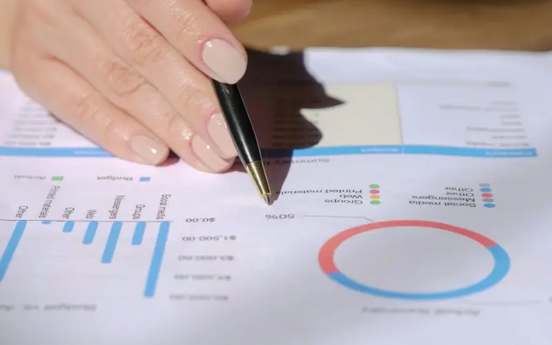 This image features a close-up of a person's hand holding a pen and pointing to a report with various data visualizations, illustrating the detailed analysis of Key Metrics for Virtual Career Fair ROI.