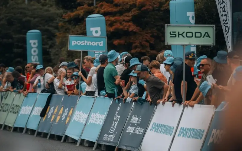 A crowd of spectators gathers behind a long row of outdoor event barrier banners displaying various corporate sponsorship logos, including PostNord, Skoda, and Sportstiming.