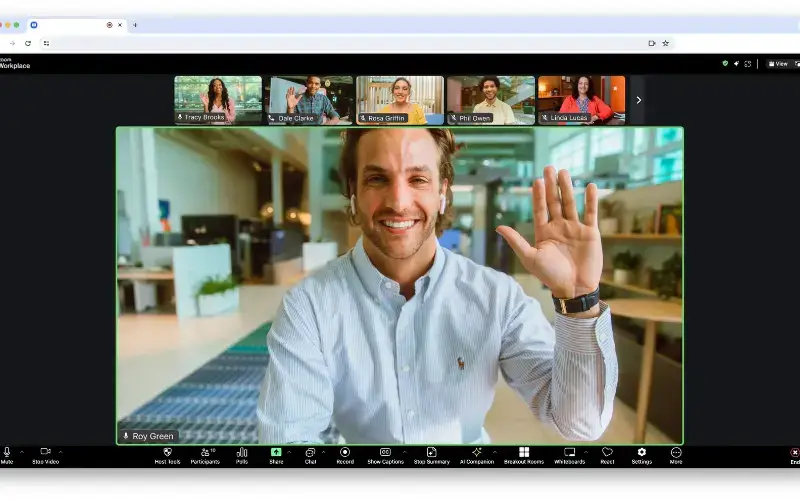 A desktop Zoom meeting interface featuring a large, central video tile of a man waving at the camera, with a row of five smaller participant video thumbnails above it.