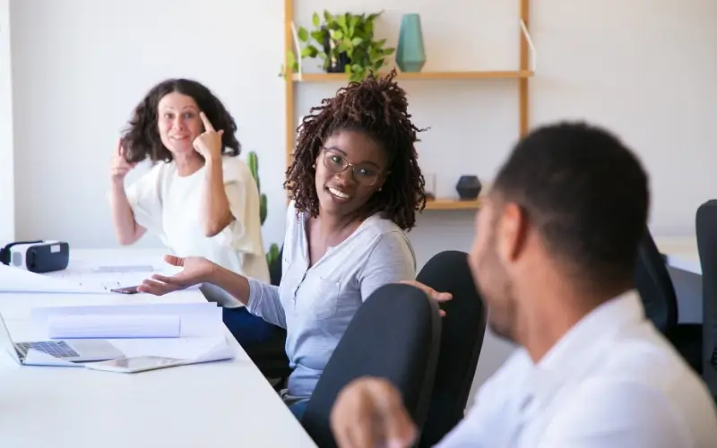 Three coworkers having an informal conversation in a bright office workspace.