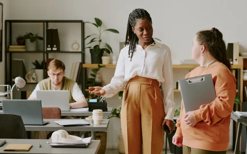 Two coworkers discussing work together in an office while another employee works nearby.
