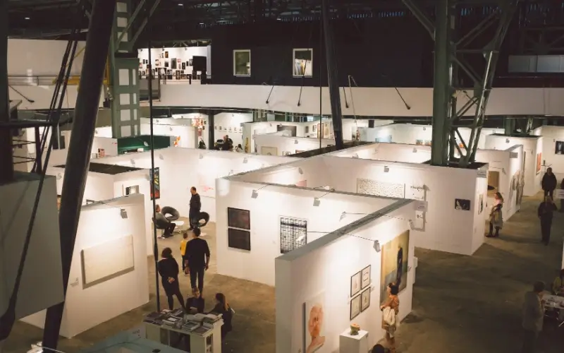 High-angle wide view of a crowded, multi-booth physical art exhibition inside a large industrial venue.