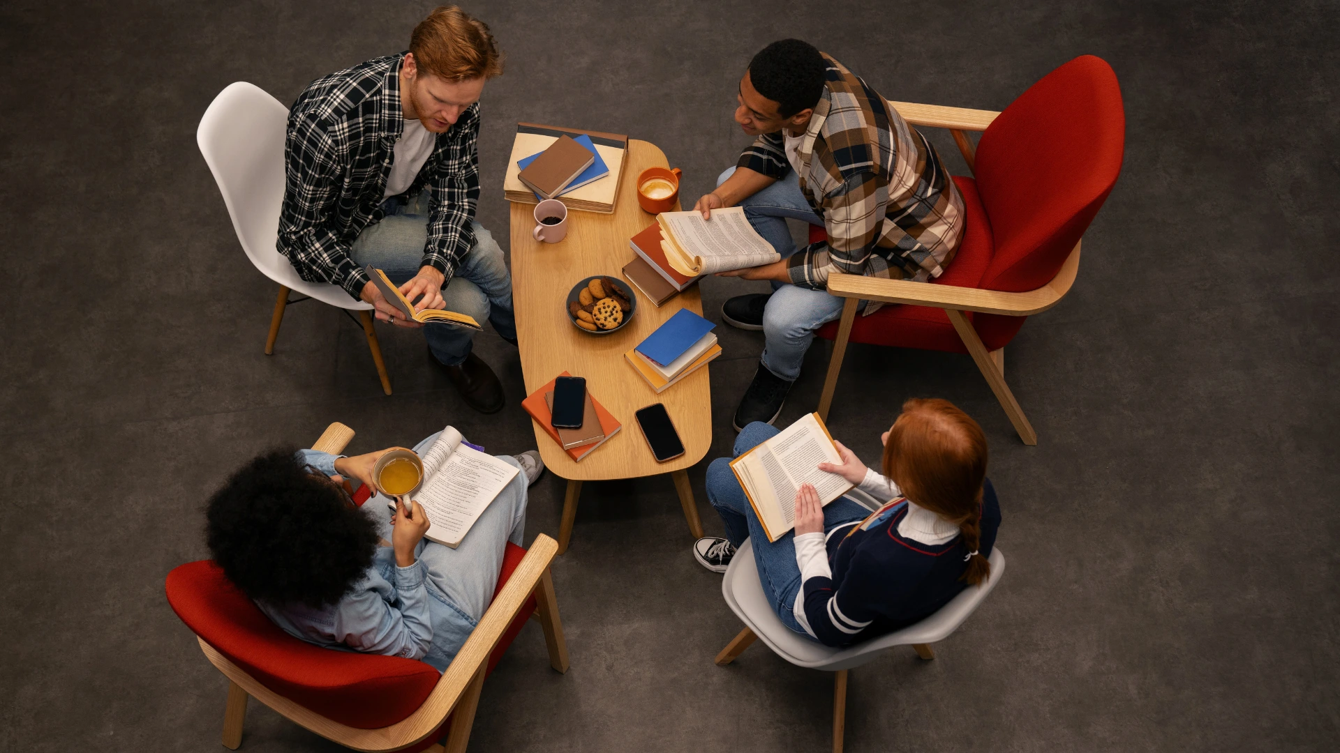 Top-down view of a diverse group of four people reading books and studying together around a coffee table in a modern lounge.