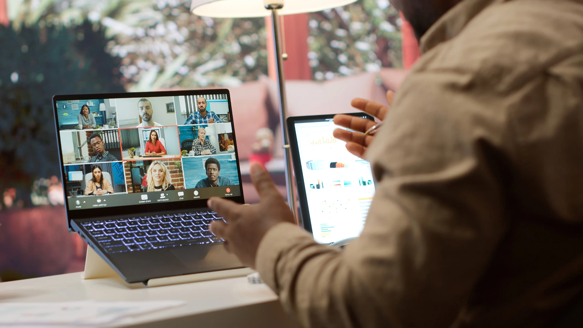 Professional reviewing business data charts on a tablet while actively speaking during a multi-person video conference call on a laptop.