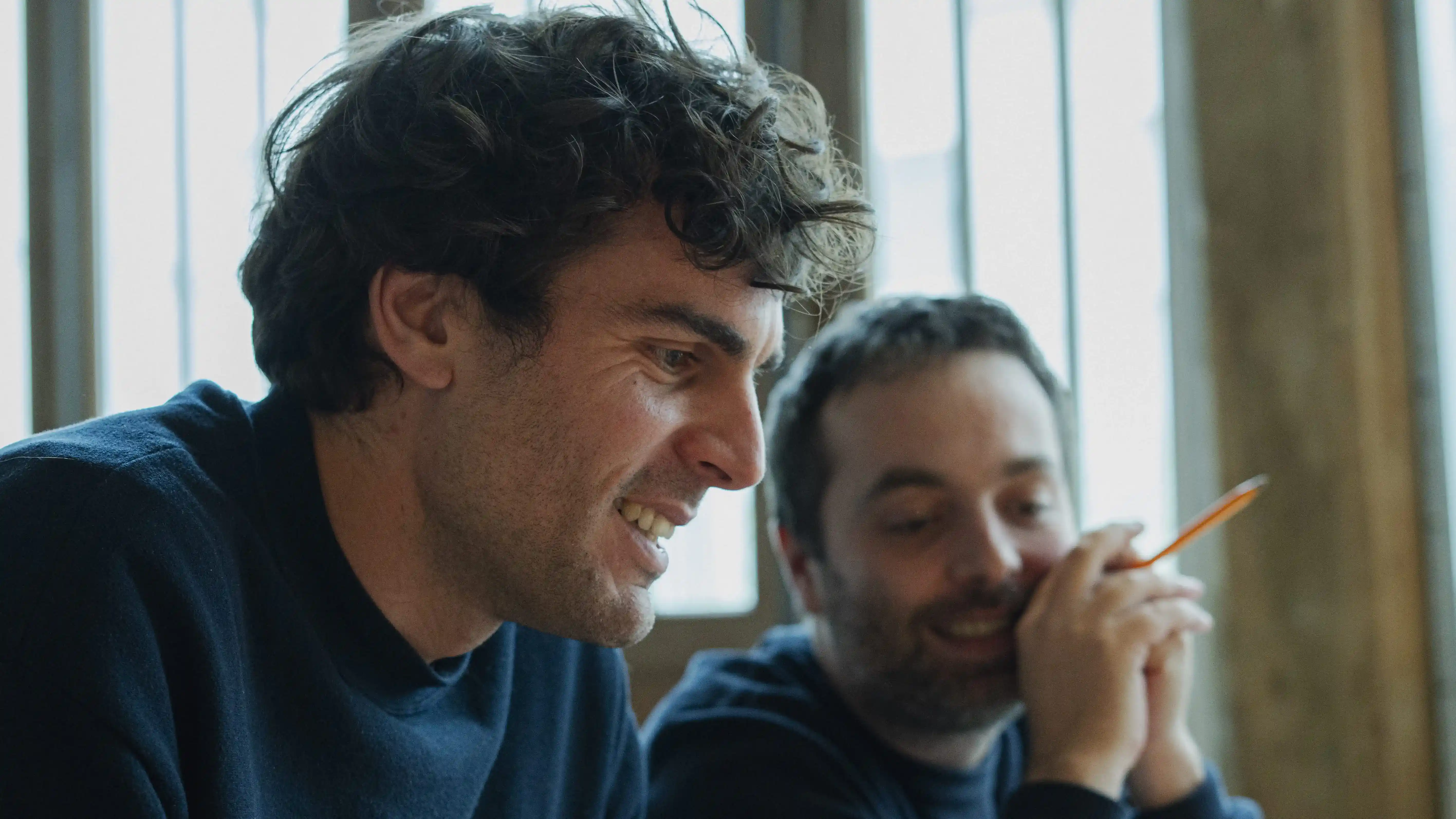 Deux collègues en pull bleu discutent près d'une fenêtre lumineuse, l'un souriant, l'autre tenant un crayon orange.