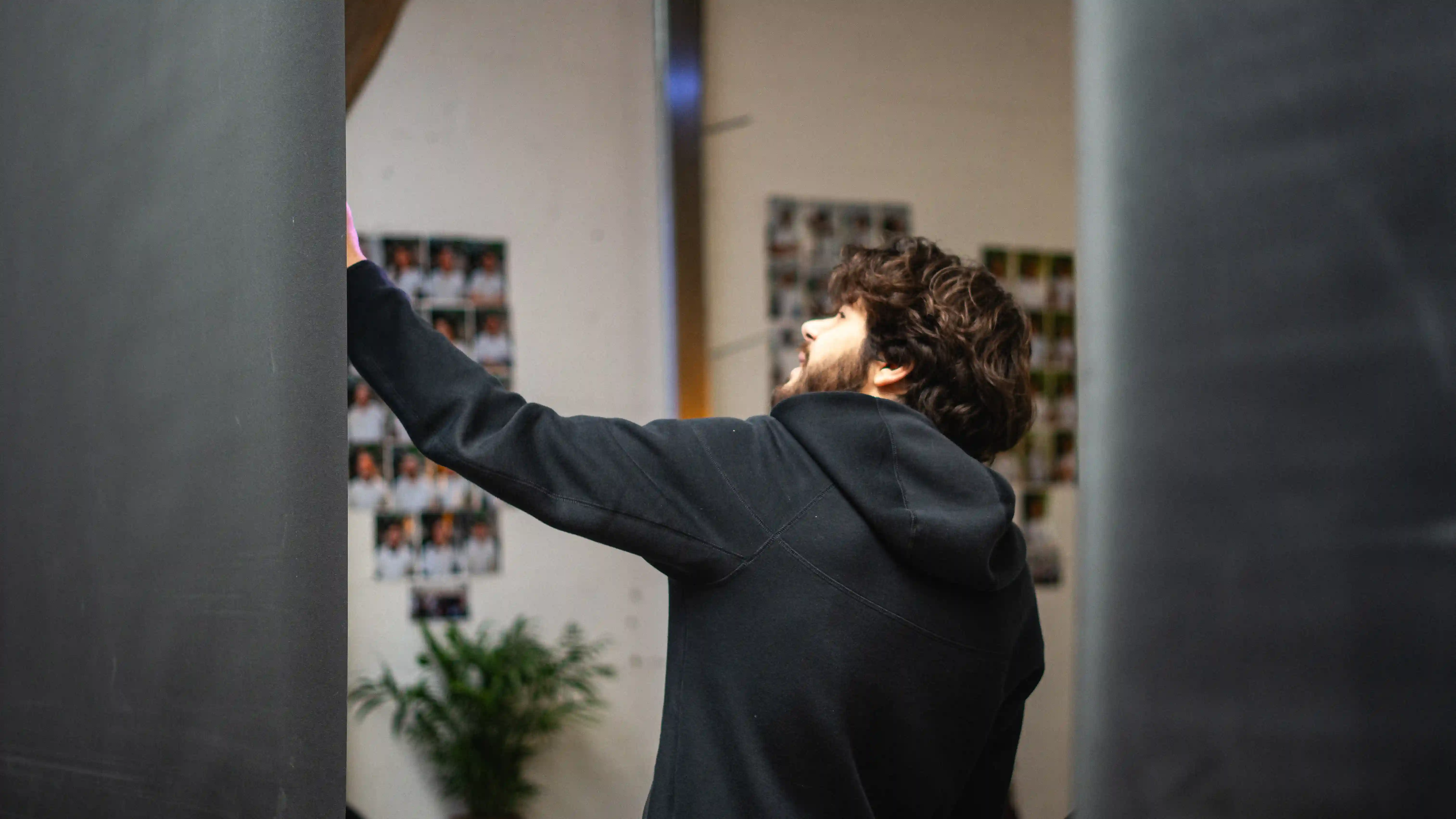 Homme barbu en hoodie sombre tend les bras vers le mur couvert de photos dans un espace intérieur.