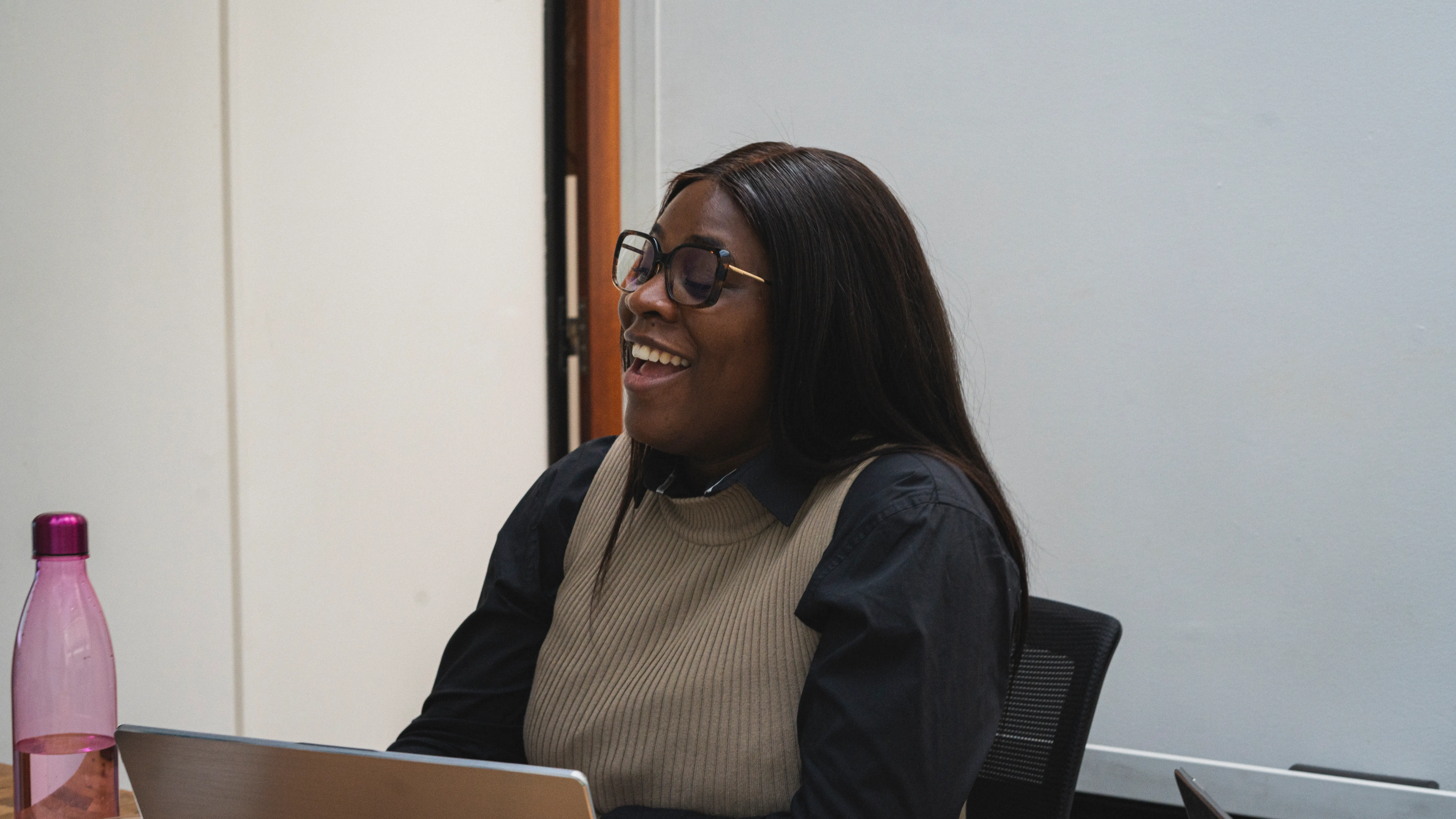 Femme souriante porte des lunettes, veston beige et chemise noire, en formation intelligence artificielle, assise devant un ordinateur.