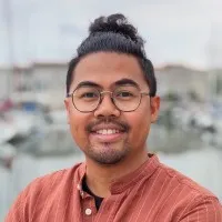 un homme à lunettes, cheveux en chignon, chemise orange, souriant sur un quai entouré de bateaux.