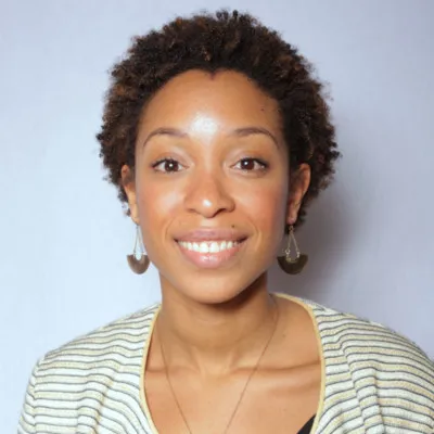 jeune femme souriante aux cheveux courts bouclés porte un pull rayé clair et des boucles d'oreilles pendantes, contre fond gris.
