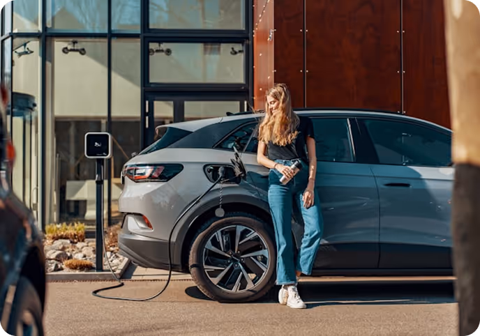 Woman standing next to a gray electric car that is charging at an outdoor charging station.