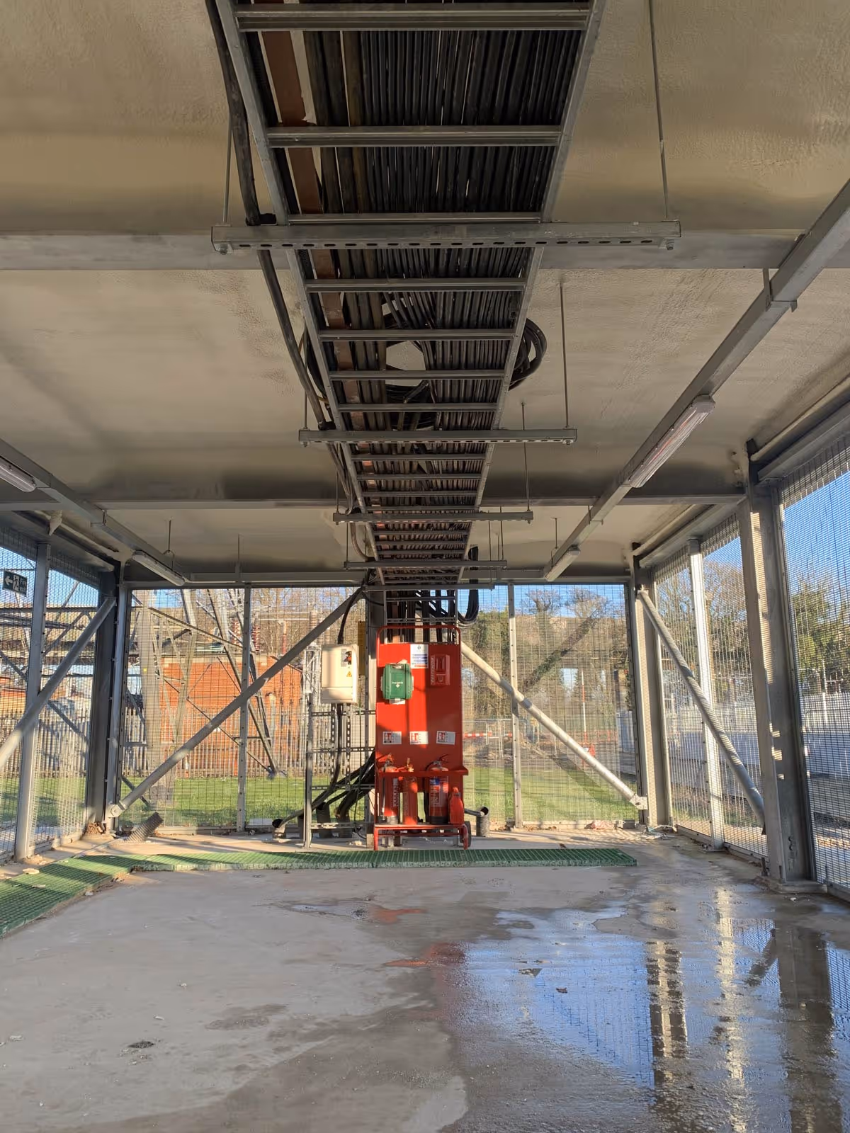 A photograph of a building structure with visible cables and a red machinery unit on a concrete floor.