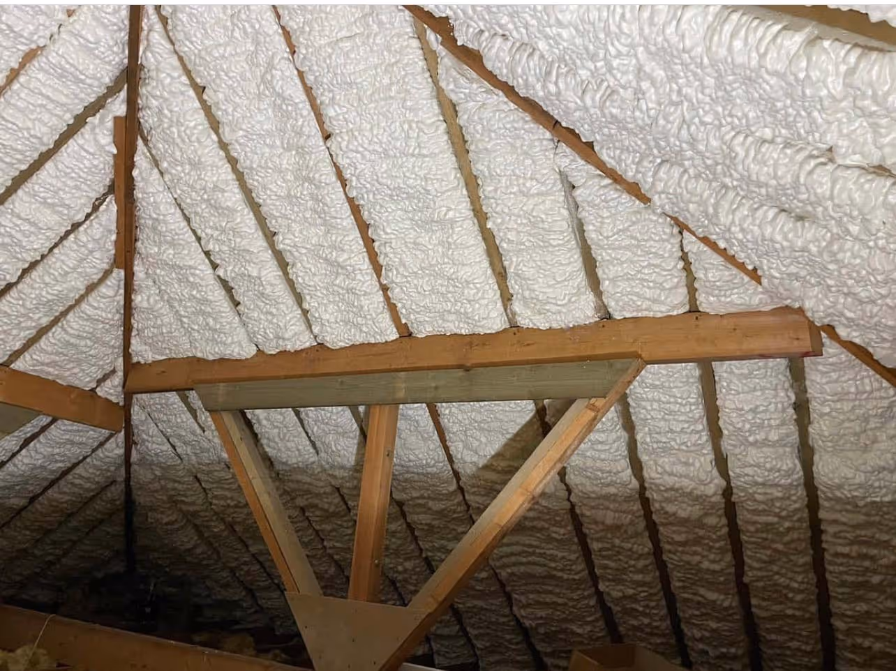 Photo of an attic roof with wooden beams and white foam insulation filling spaces between rafters.