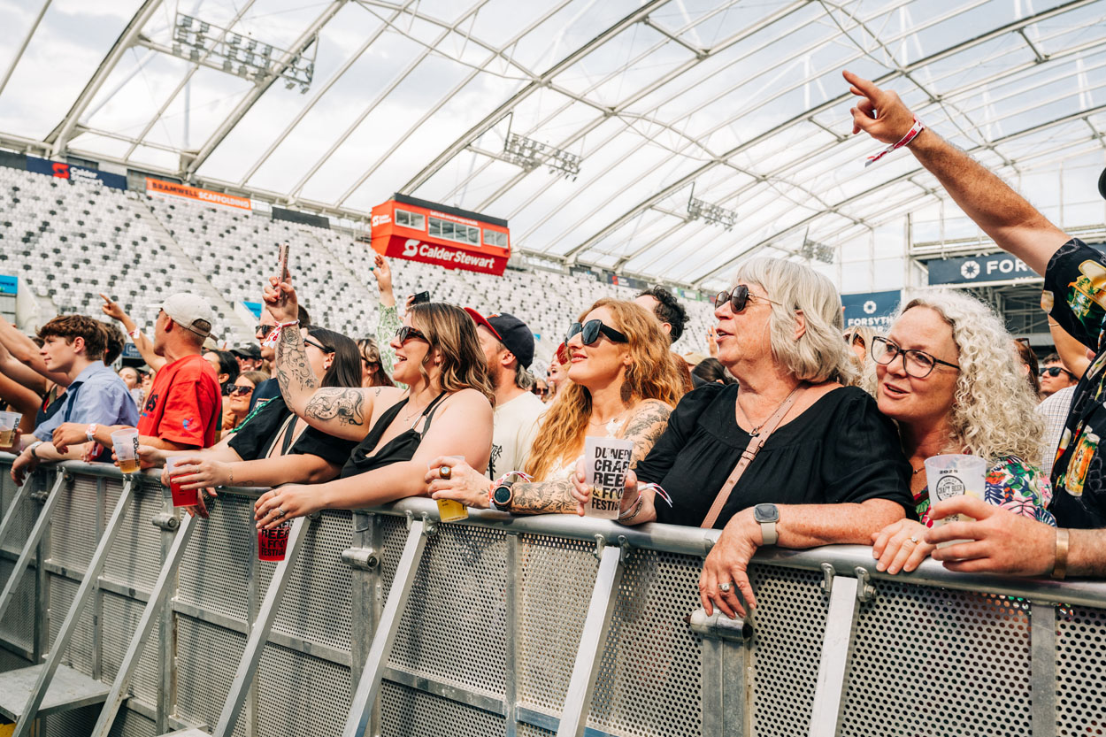 Crowd enjoying a music festival inside a stadium, some holding drinks and phones near the front barrier.