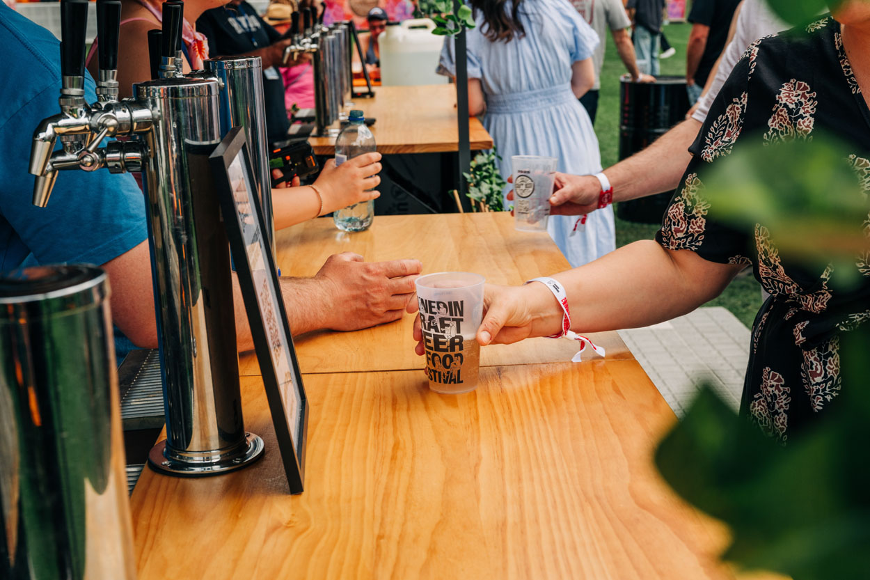 Person holding a craft beer cup at a festival bar with taps and people in background.