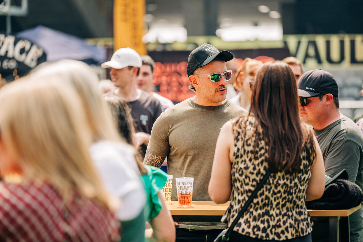 Group of people chatting at an outdoor festival with craft beer cups on a wooden table.
