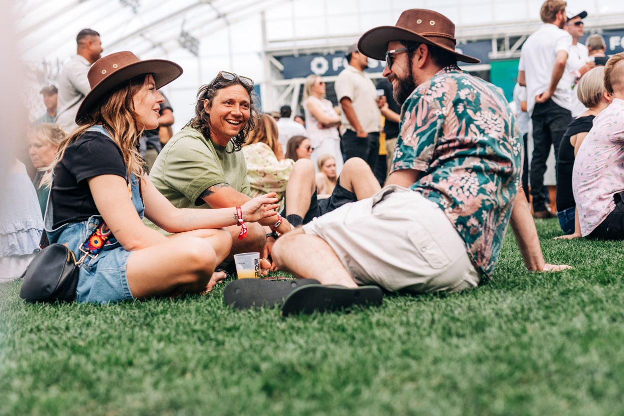Three people sitting on grass chatting at a crowded outdoor event under a large tent.