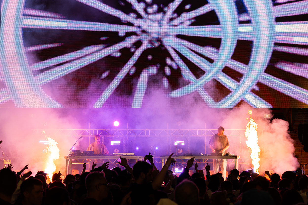 Electronic music DJs performing on stage with flames, lights, and a vibrant digital backdrop at concert.