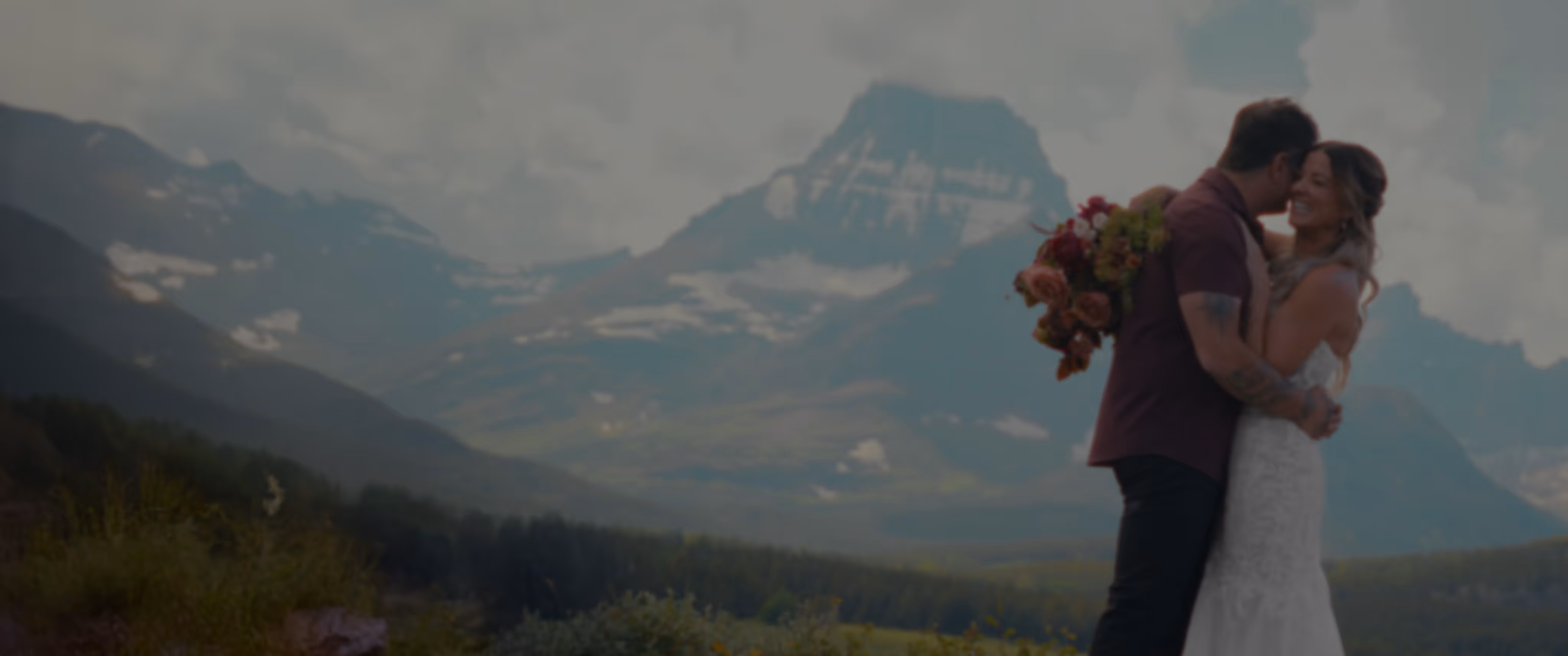 Bride and groom embracing outdoors with mountainous landscape and partly cloudy sky in the background.
