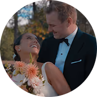 Groom in dark suit tenderly embraces bride in white dress holding a bouquet, both outdoors among trees.