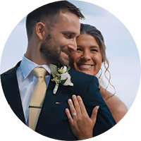Groom in dark suit tenderly embraces bride in white dress holding a bouquet, both outdoors among trees.