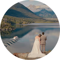 Groom in dark suit tenderly embraces bride in white dress holding a bouquet, both outdoors among trees.