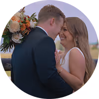 Groom in dark suit tenderly embraces bride in white dress holding a bouquet, both outdoors among trees.