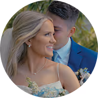 Groom in dark suit tenderly embraces bride in white dress holding a bouquet, both outdoors among trees.