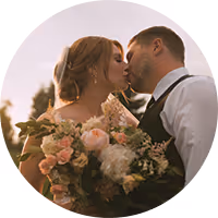Groom in dark suit tenderly embraces bride in white dress holding a bouquet, both outdoors among trees.