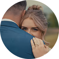 Groom in dark suit tenderly embraces bride in white dress holding a bouquet, both outdoors among trees.