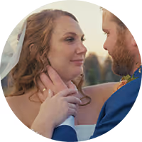 Groom in dark suit tenderly embraces bride in white dress holding a bouquet, both outdoors among trees.