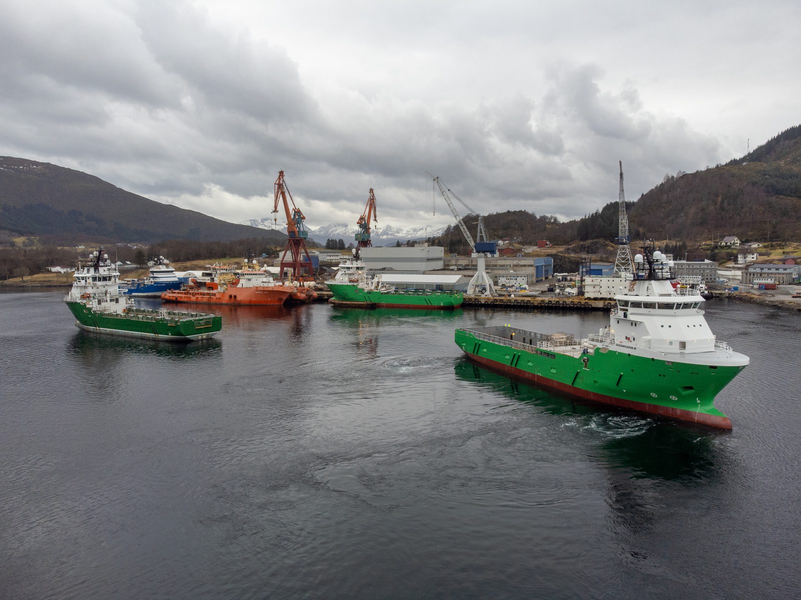 Oceanicasub IX underway near a shipyard quay with sister vessels and support craft moored behind it, snow-capped mountains beyond