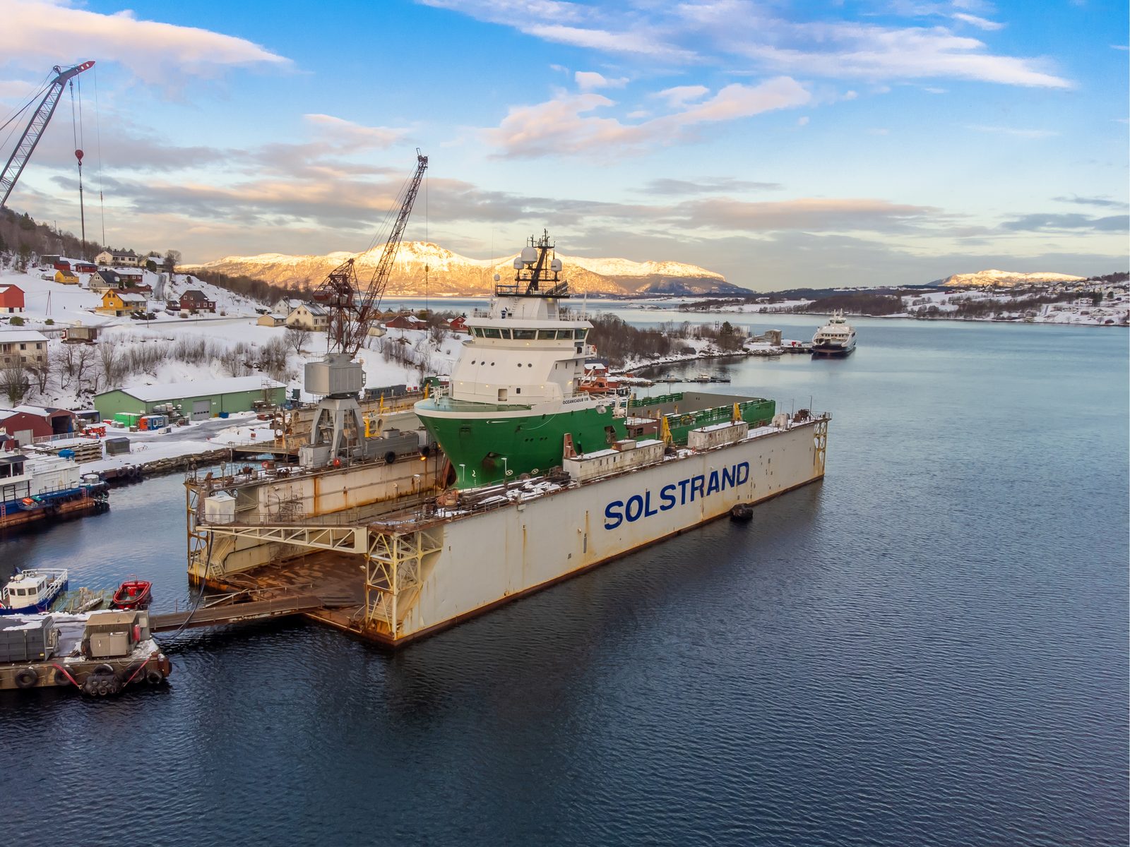 Oceanicasub VIII in the Solstrand floating dry dock at golden hour, with snow-capped mountains and a ferry in the background fjord