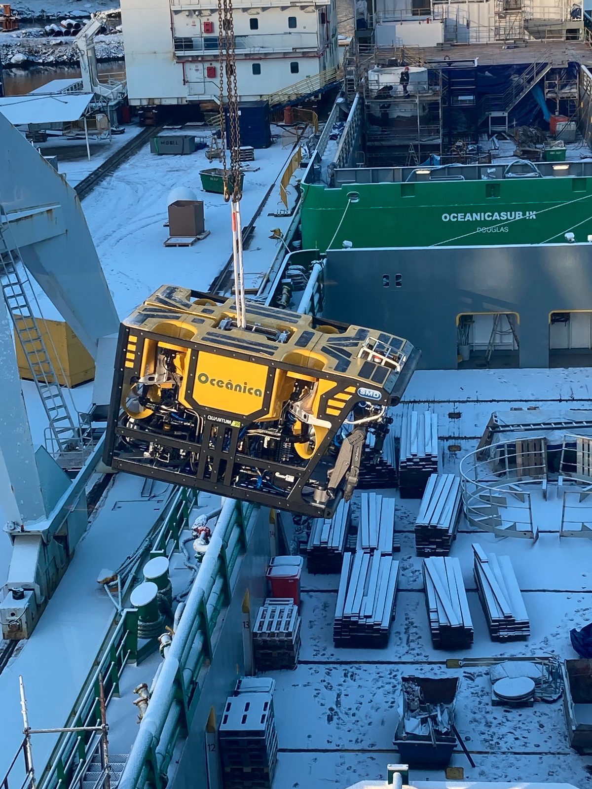 Yellow Oceânica-branded work-class ROV being lifted by crane onto a snow-covered vessel deck, with Oceanicasub IX and stacked steel plate visible below.