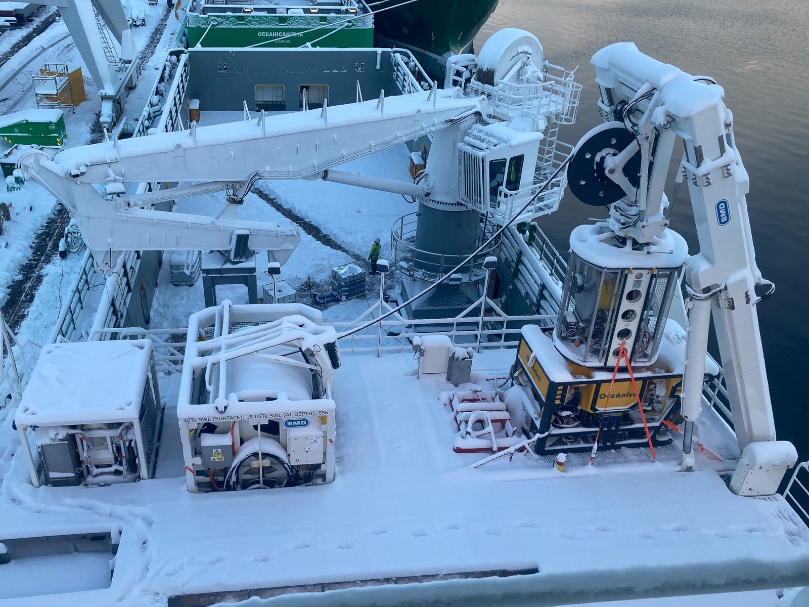 Snow-covered offshore support vessel deck with ROV launch-and-recovery winch and a yellow Oceânica work-class ROV secured on board; sister vessel Oceanicasub IX moored alongside.
