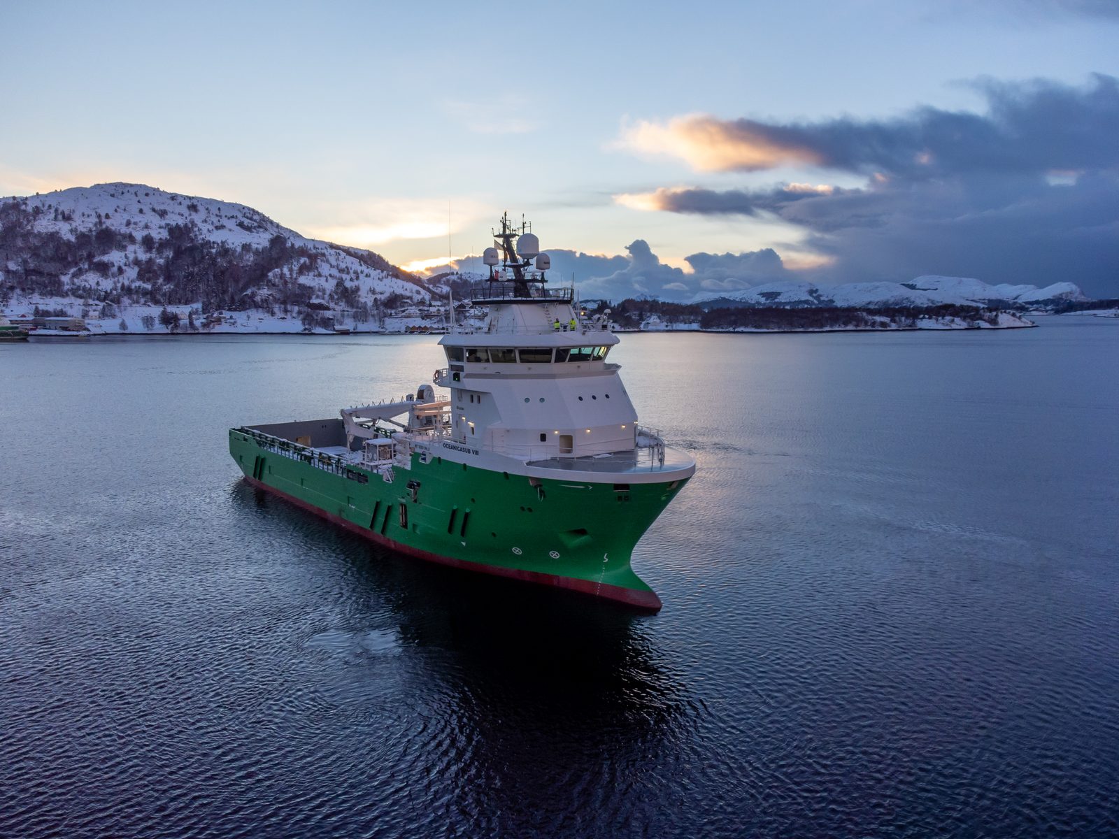 Oceanica offshore support vessel underway on calm water at dusk, snow-covered hills and pastel sunset sky, two crew visible on the bridge wing.