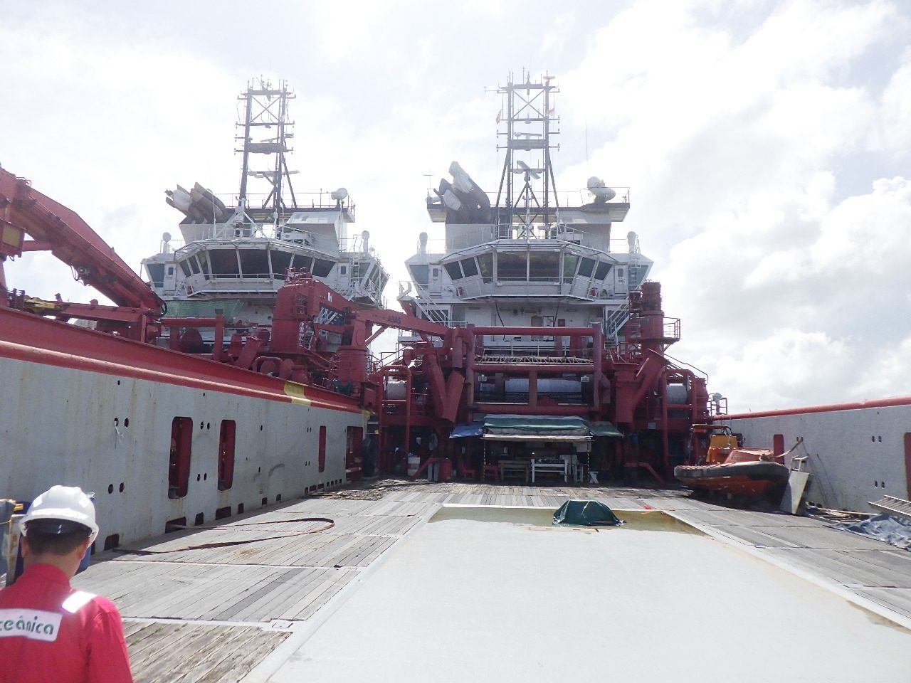 View aft from an AHTS deck toward a sister vessel moored alongside, red offshore handling cranes visible, an Oceânica crew member in the foreground