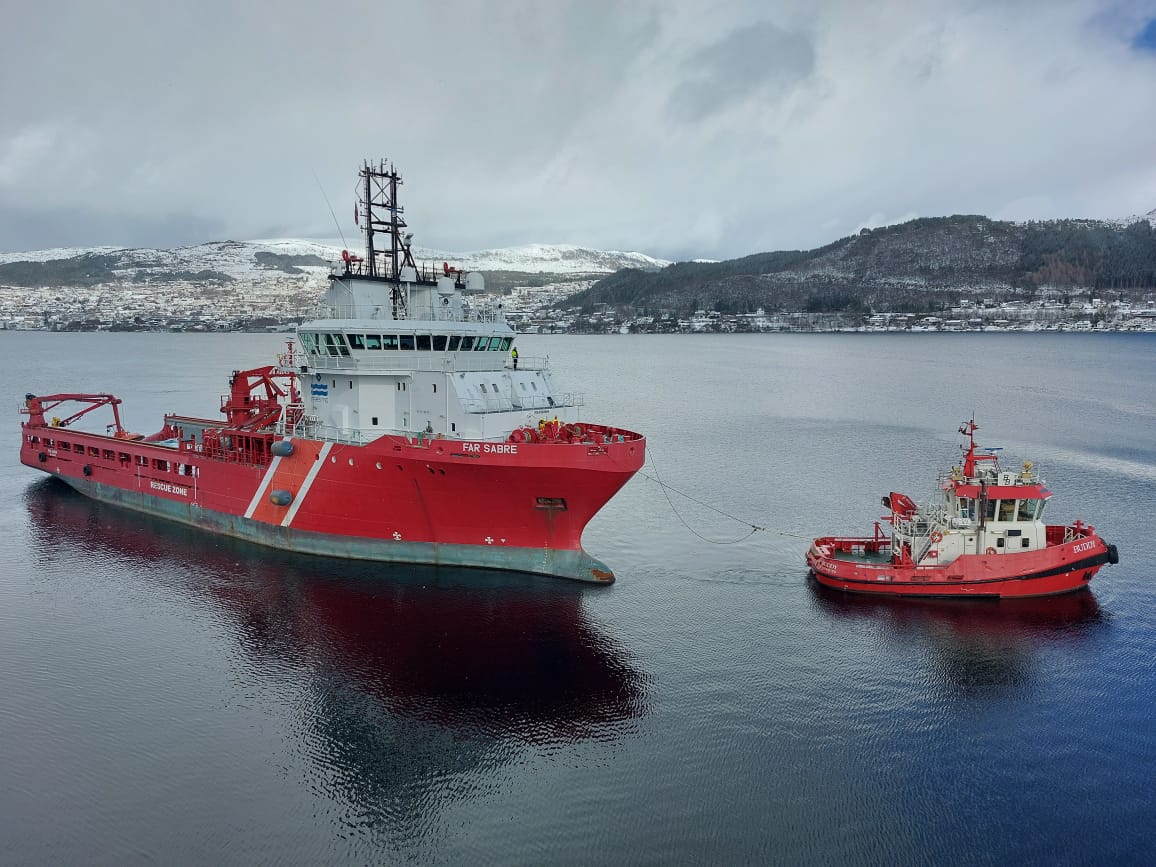 Aerial view of Far Sabre under tow by a harbour tug across calm fjord water, snow-covered hills in the background
