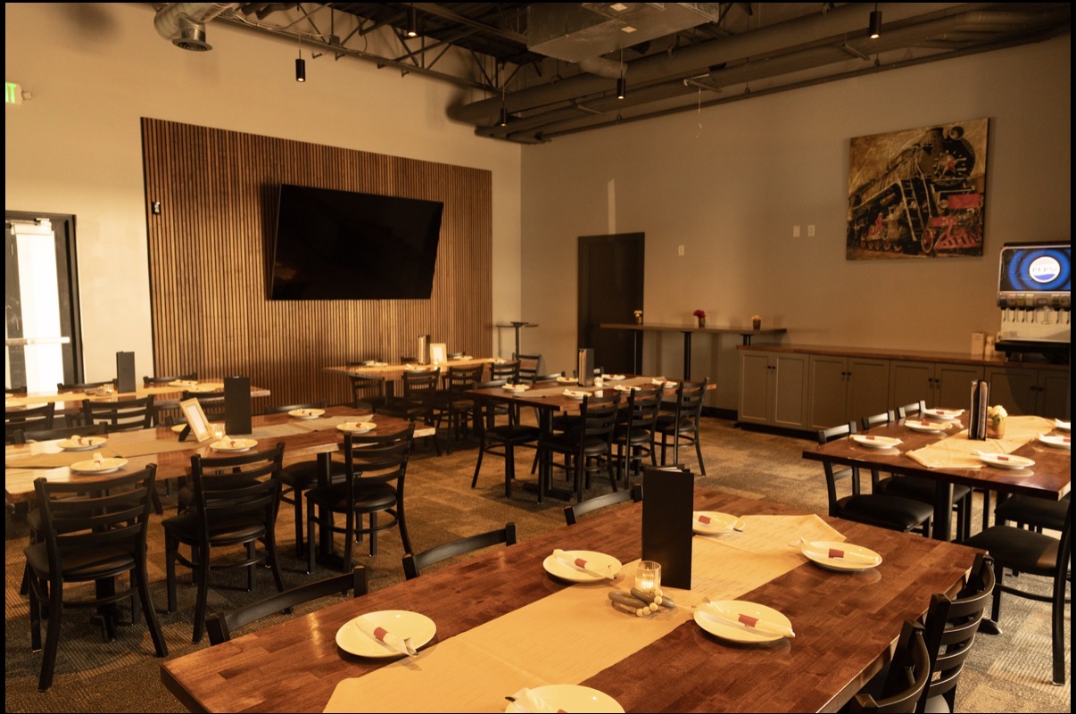 Warmly lit dining area with wooden tables and black chairs set with white plates, napkins, utensils, and candles, featuring a large wall-mounted TV, wood panel accent wall, beverage dispenser, and a painting of a train on a beige wall.