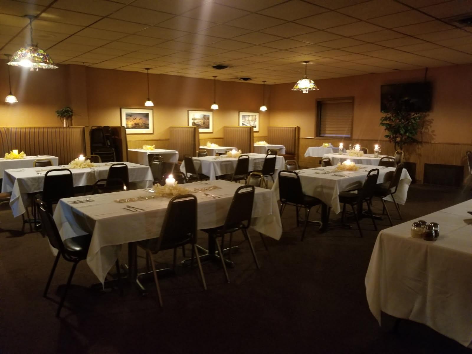 Dimly lit banquet room with several rectangular tables covered in white tablecloths, each with candles and floral centerpieces, surrounded by black chairs, and booth seating along the walls under hanging lights and framed pictures.