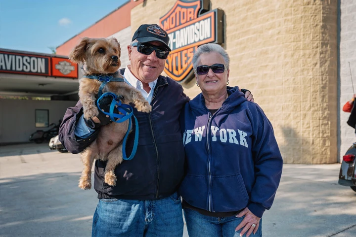 Senior couple holding Yorkie Terrier posing in front of Harley-Davidson signage