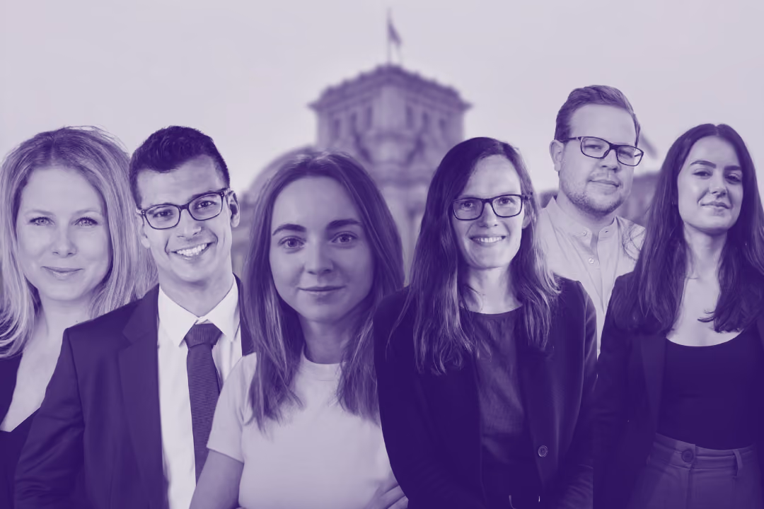 Eine Gruppe von sechs lächelnden jungen Erwachsenen steht im Vordergrund vor dem unscharf abgebildeten Reichstagsgebäude in Berlin.