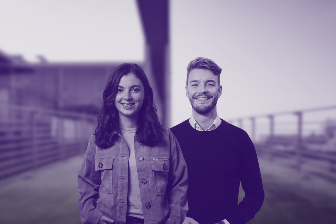 Zwei lächelnde junge Erwachsene stehen nebeneinander vor einem unscharfen Außenhintergrund.